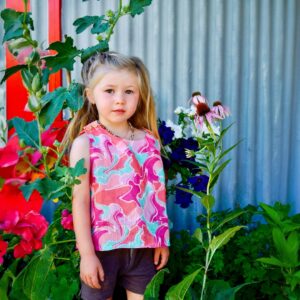 Blonde girl with long hair is standing at front of metal fence. She is surrounded with flowers. Wearing Girls crop top abstract with pastel print in pinks and aqua- original textile design by Miramara