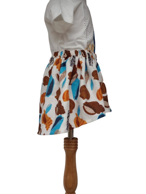 Mia Skirt 1 is uneven length girl's skirt which is displayed on a mannequin. The print shows different colored seashells in brown, blues, orange and beige.