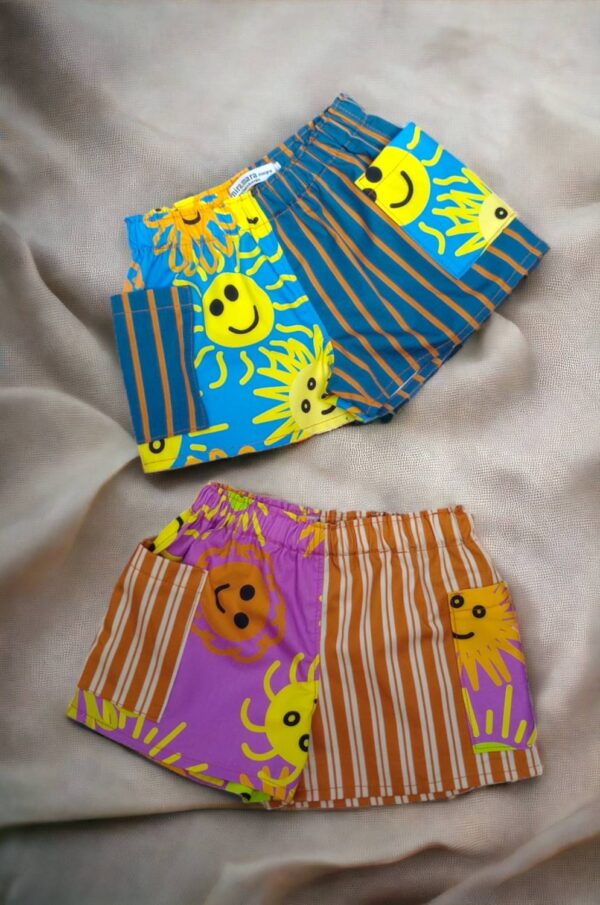 A pair of Alana kid's shorts over crinkled fabric. One pair is in blue stripes with orange. And the other using orange and cream stripes. Opposite print is featuring sun faces printed on blue or purple background.
