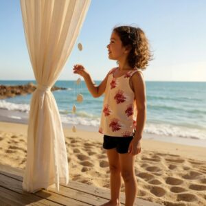 Child wearing sleeveless pink lionfish print top and dark indigo shorts from Clothing Bundle 4, standing on sandy beach near ocean, holding decorative hanging shells beside sheer white curtain in breeze.